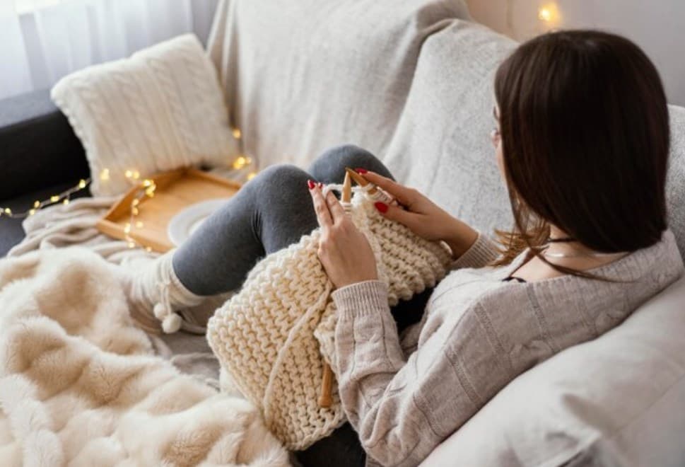 a woman using knitting tools to knit a lap blanket while sitting on a couch