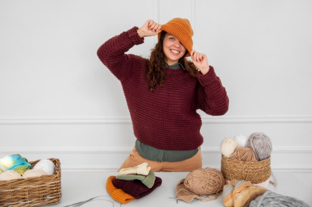 A woman adjusts an orange knitted hat on her head, surrounded by skeins of yarn and knitted items