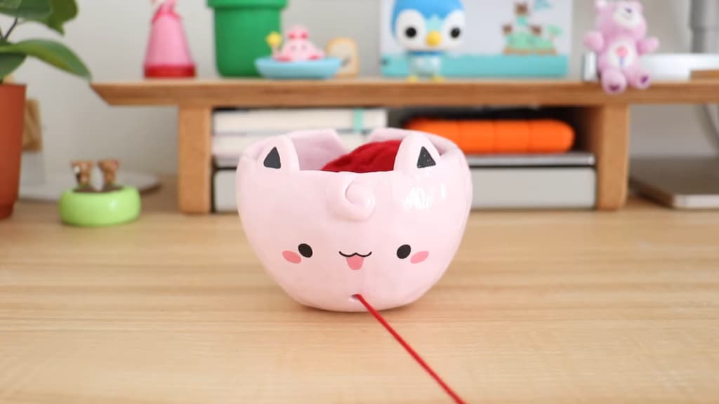 A cute pink cat-shaped yarn bowl on a wood table with plush toys in the background