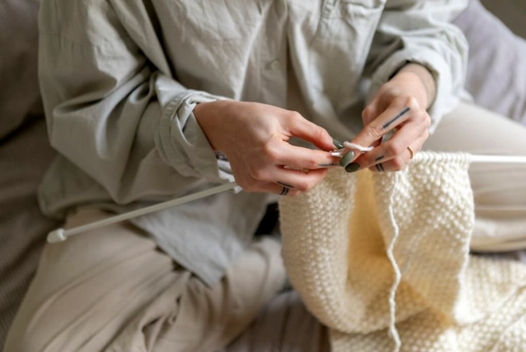 Close-up of hands knitting a cream-colored fabric with white knitting needles