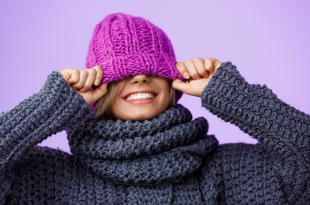 A smiling woman pulls a purple knitted hat over her eyes, wearing grey knitted scarf