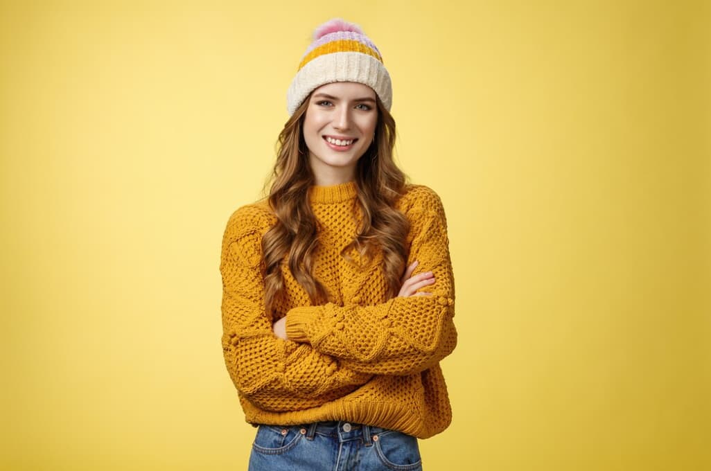 A smiling woman with long hair wears a mustard yellow knit sweater against a yellow background