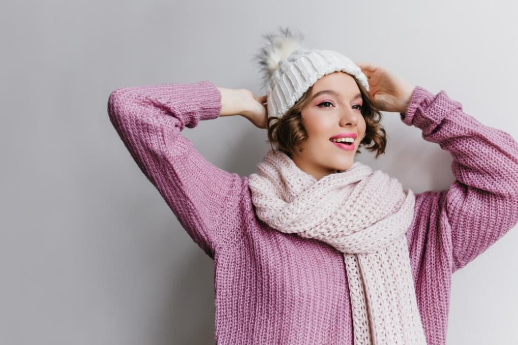 A woman with short hair dressed in a chunky pink knit sweater and scarf against a white wall