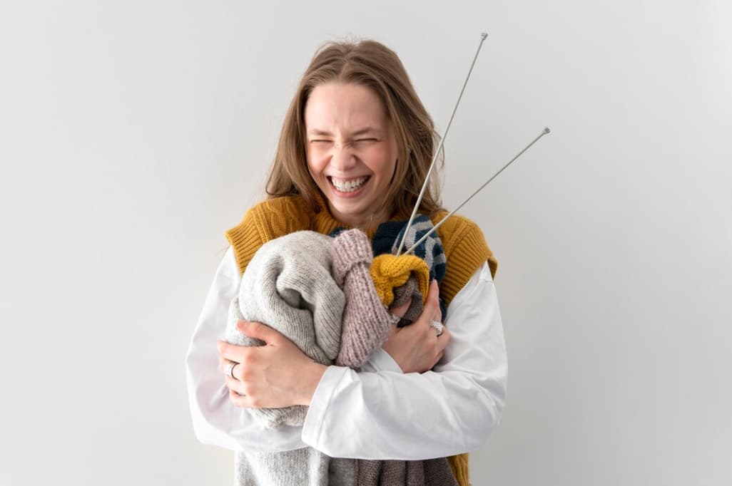 Joyful woman hugging knitted clothes with knitting needles on white background